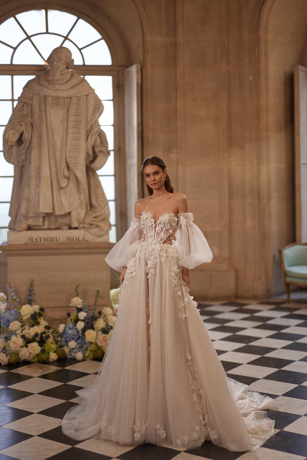 Robe de mariée Celestine, avec un décolleté ouvert, des manches légères laissant les épaules dénudées et des fleurs en relief — collection Something Blue 2027, DAMA Couture.