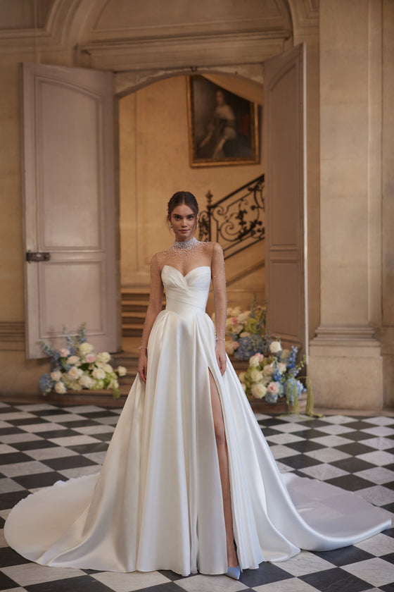 Robe de mariée trapèze Lucienne, avec un décolleté illusion orné de cristaux et une jupe drapée en satin avec une fente haute.
