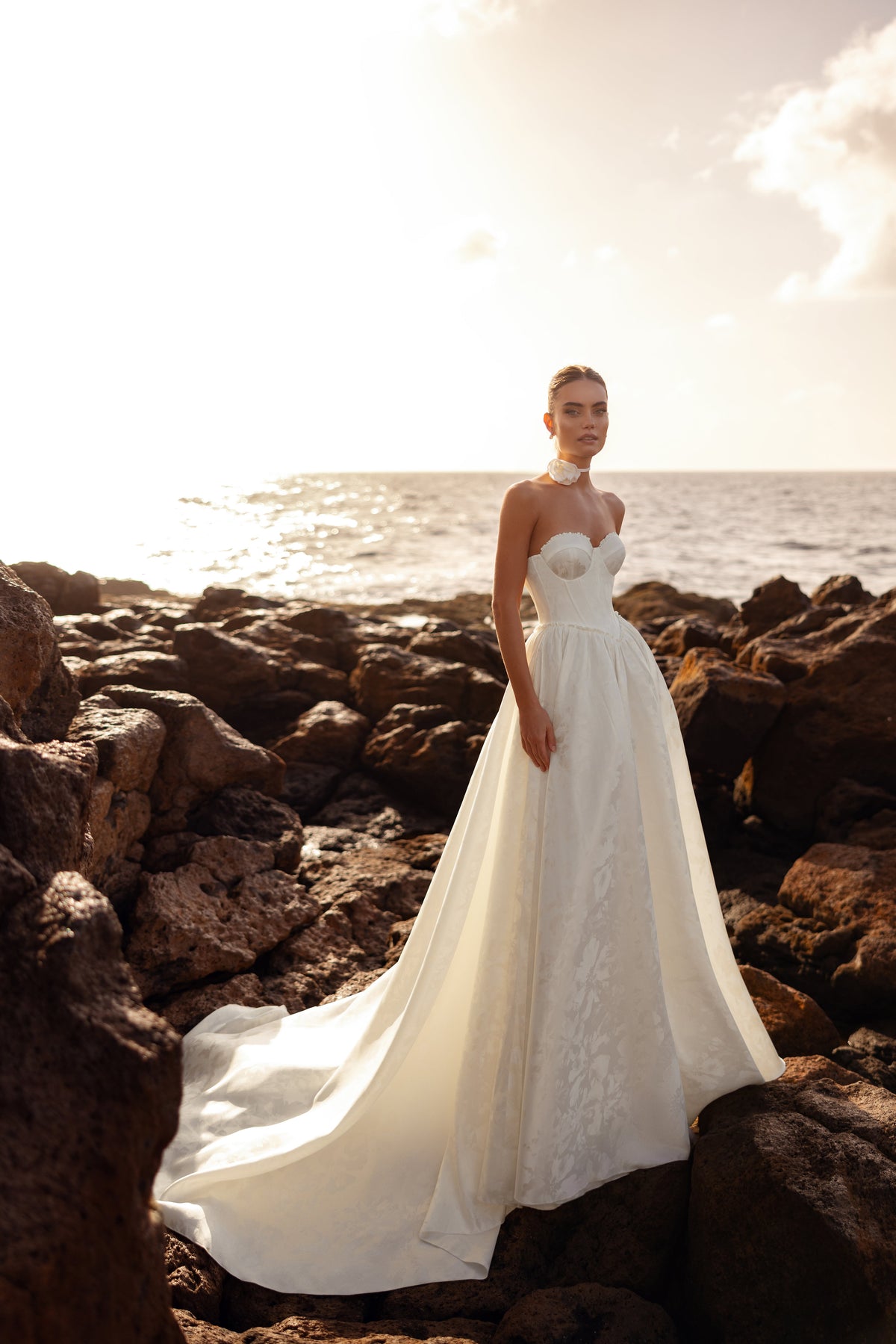 Robe de mariée Cholly, avec un bustier structuré et une longue traîne, au coucher du soleil au bord de la mer.