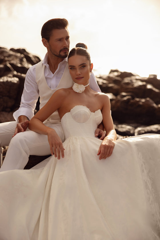 Robe de mariée Cholly au fini satiné, avec un corsage structuré et une jupe ample, photographiée au bord de la mer.