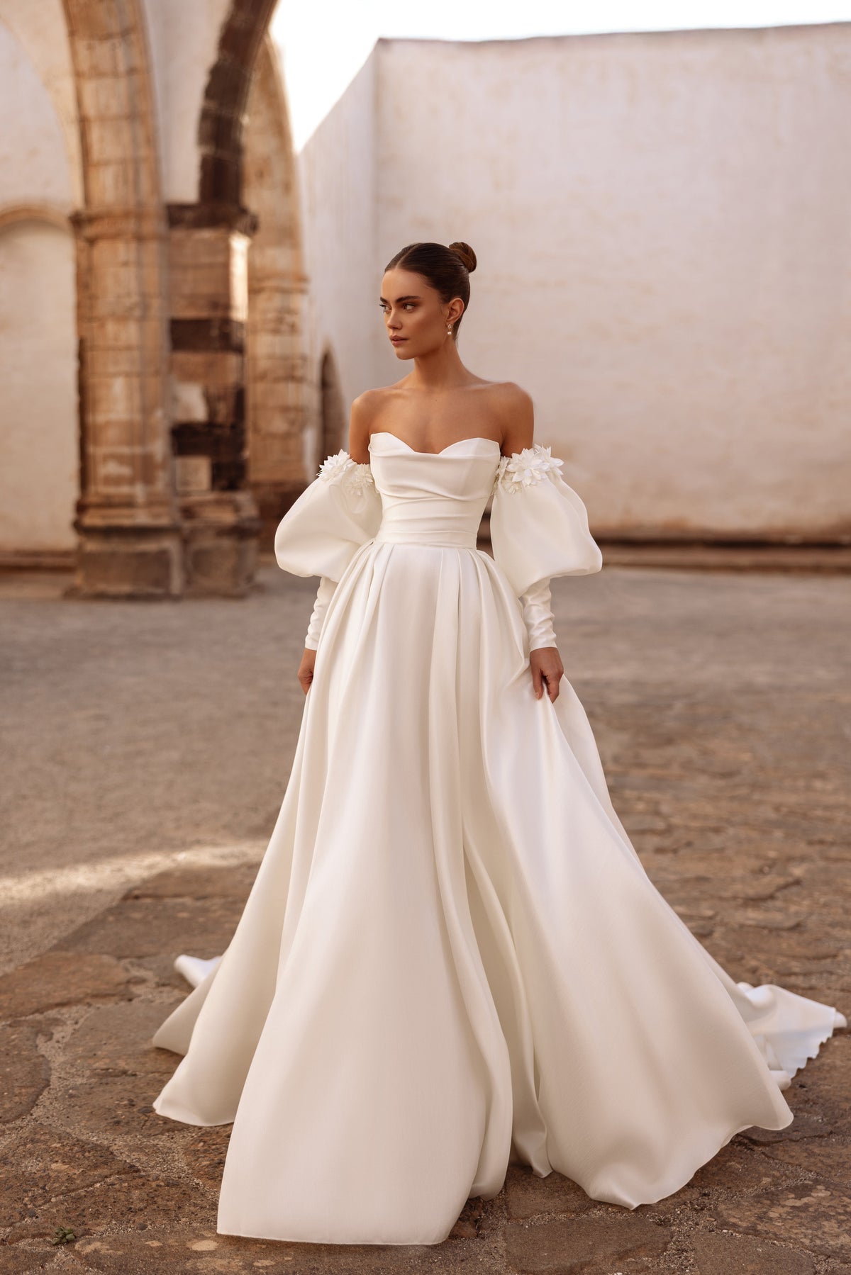 Robe de mariée Anika en satin, avec des épaules dénudées, des manches bouffantes et des appliqués floraux.