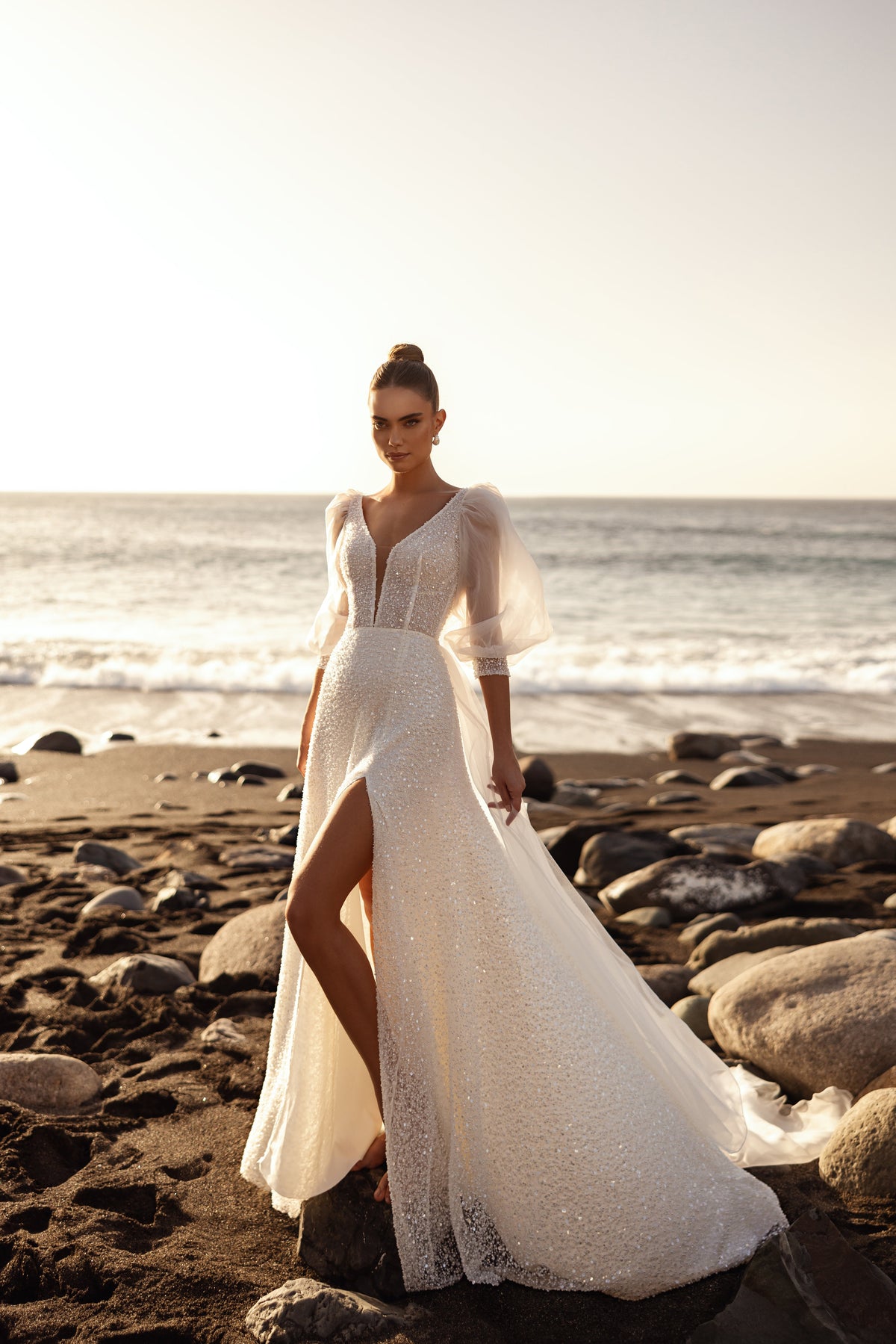 Robe de mariée Davina, avec un décolleté en V plongeant et un tissu scintillant au coucher du soleil sur la plage.