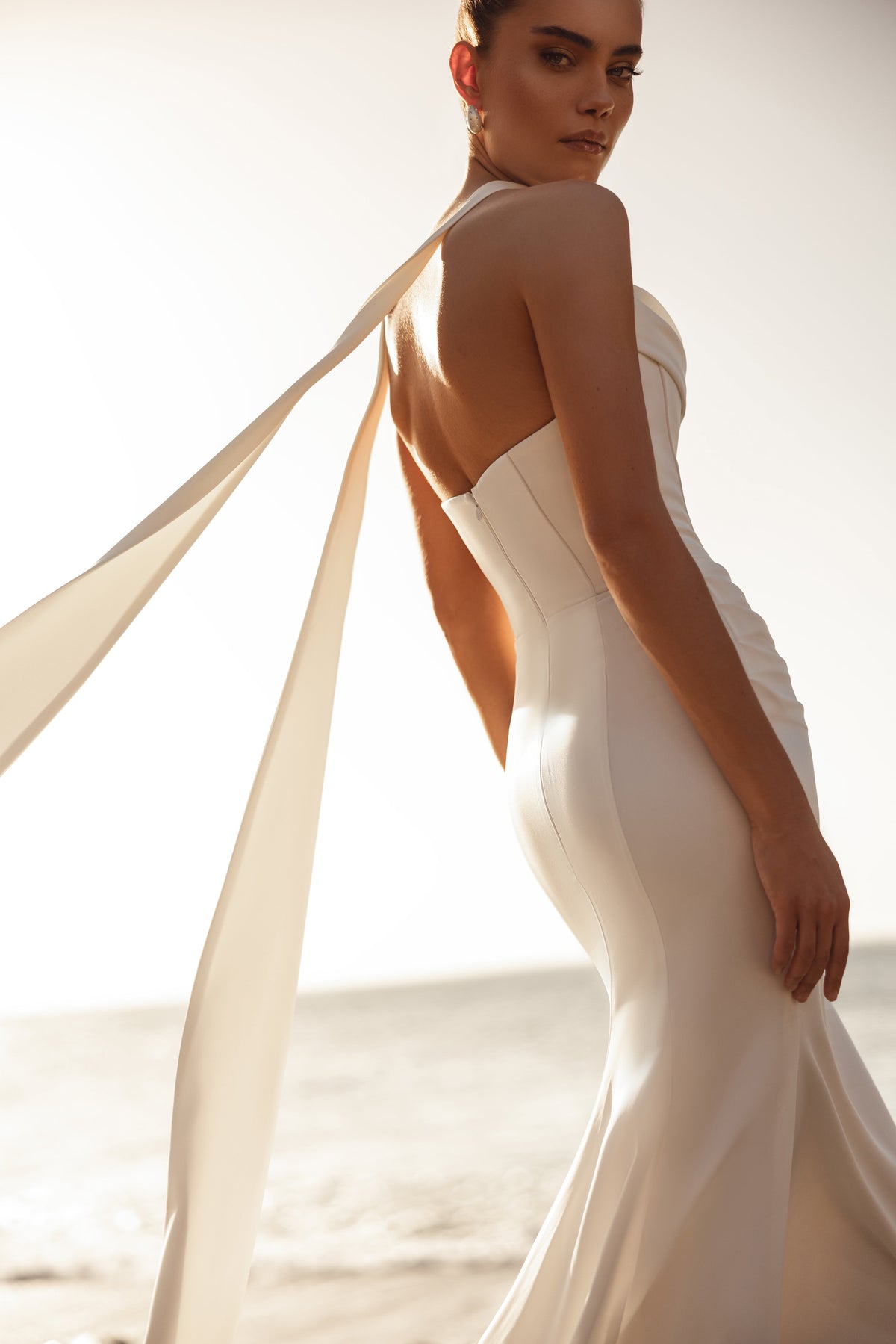 Robe de mariée Luna, avec une longue traîne et des rubans flottants, au bord de l’océan.