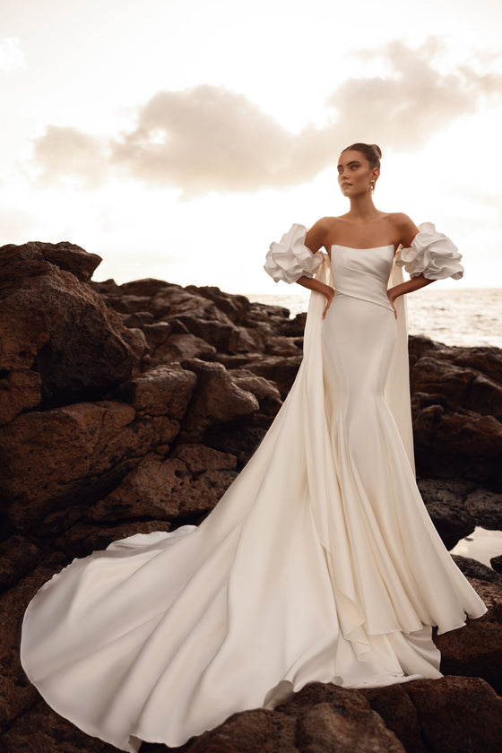 Robe de mariée Soraya, avec des manches bouffantes et une longue traîne, au bord de la mer.